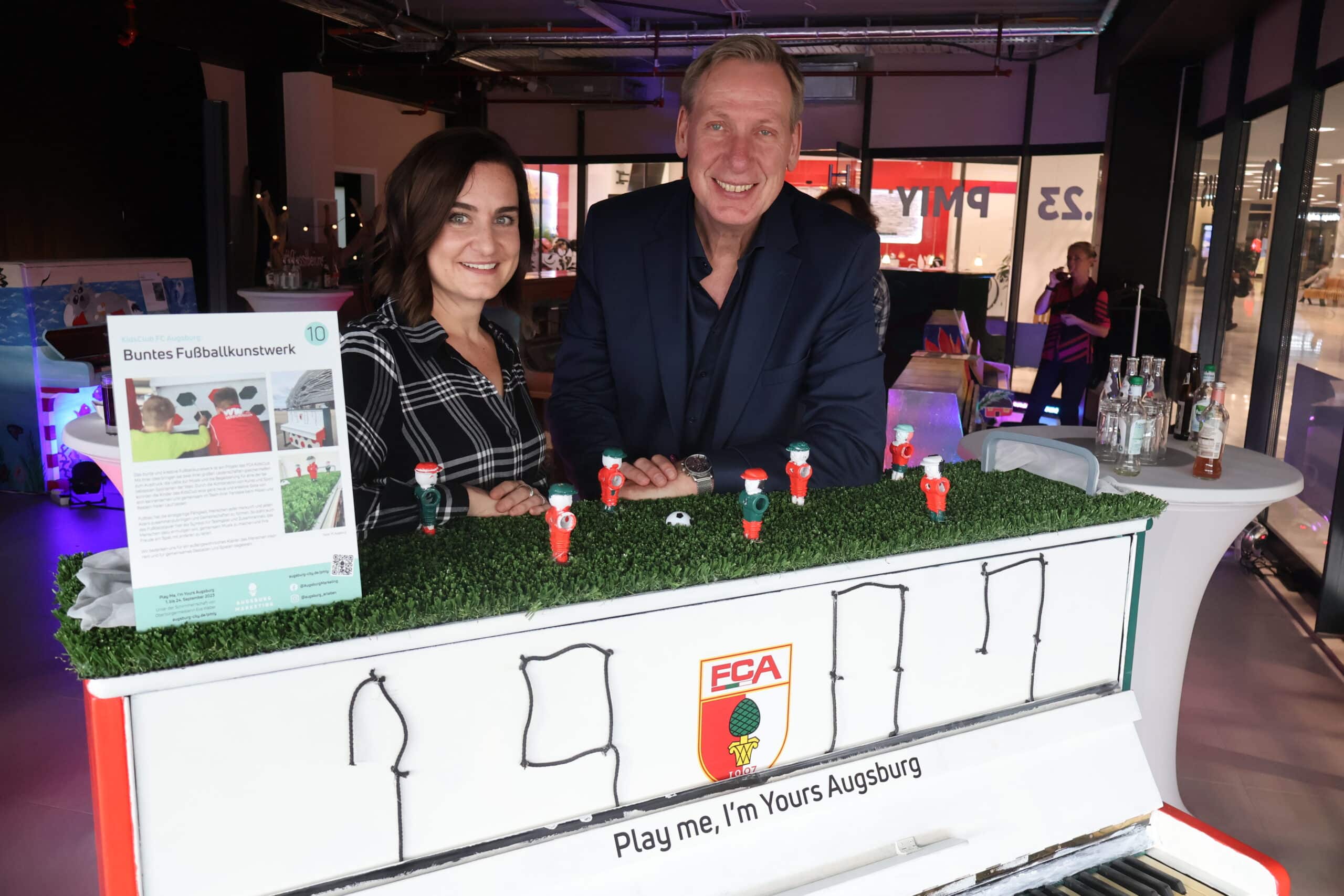 Annemarie Cizmadia und Heinz Stinglwagner vom Augsburg Marketing hinter dem „Bunten Fußballkunstwerk“, das der FCA KidsClub gestaltet hat. Es steht in der Innenstadt auf dem Martin-Luther-Platz.