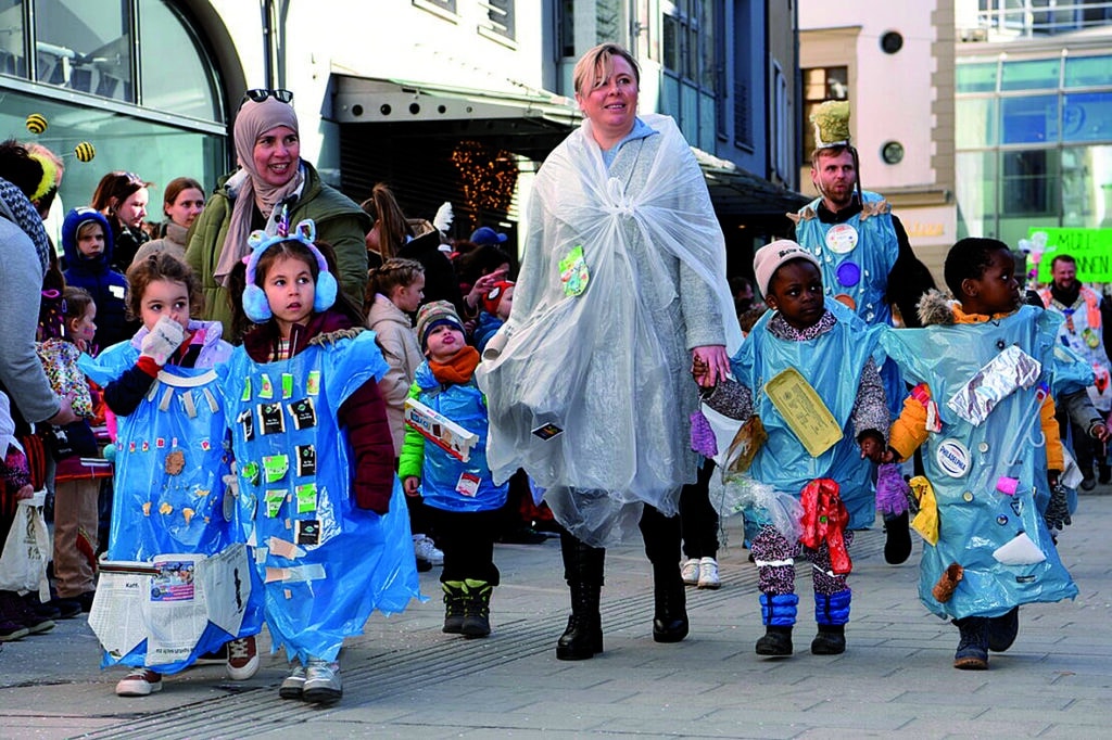 Auf dem Bild sieht man den Gaudiwurm in der Innenstadt: Kinder in blauen und eine Betreuerin in einem weißen Kostüm.